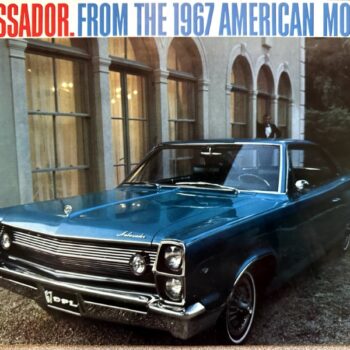 Front three-quarter view of a blue 1967 American Motors Ambassador parked in front of a stone building with arched windows.