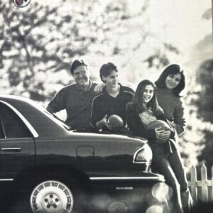 Group of four young adults leaning on a dark Buick LeSabre, 1999 model, smiling outdoors with a picket fence in the background and soft bokeh light.