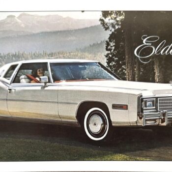 Vintage Cadillac Eldorado coupe parked on a scenic overlook with mountains in the background; a luxuriously styled white car facing left, driver visible through the window.