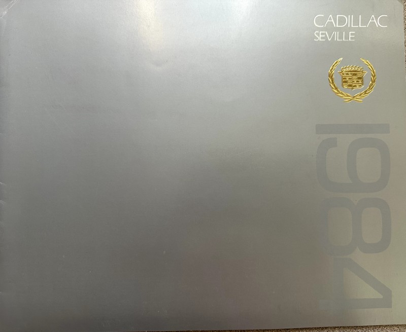 IMG_5805 | Merrick Auto Museum Metallic gray Cadillac Seville brochure cover with the white 'CADILLAC SEVILLE' text at the top right and a gold crest; large faded numbers run vertically along the right edge.