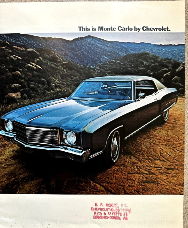 IMG_5887 | Merrick Auto Museum Blue vintage Chevrolet Monte Carlo parked on a dirt road with rocky mountains in background; advert text above reads 'This is Monte Carlo by Chevrolet.'