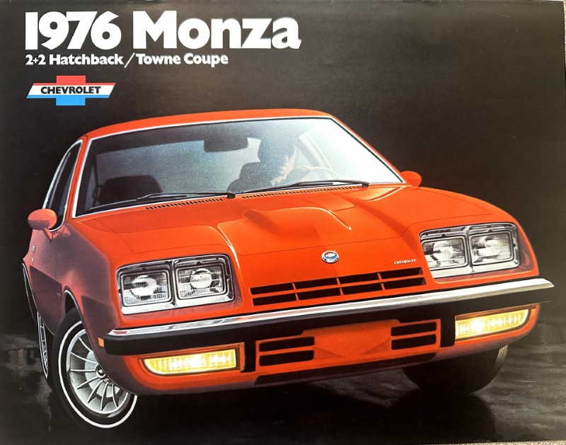 IMG_5986 | Merrick Auto Museum Front view of a bright orange 1976 Chevrolet Monza 2+2 hatchback in an ad cover against a dark background, headlights on.