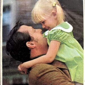 Man lifting a young girl in a green dress, both smiling at each other outdoors; nostalgic 1969 Pontiac promo photo.