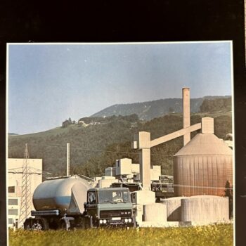 Industrial plant with silos and a DAF 2300 truck in a field of yellow flowers, hills in the background.