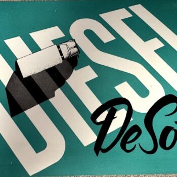 Teal sign with large diagonal white 'DIESEL' text and black cursive 'DeSoto' overlay; a white‑and‑black object rests on the letters.