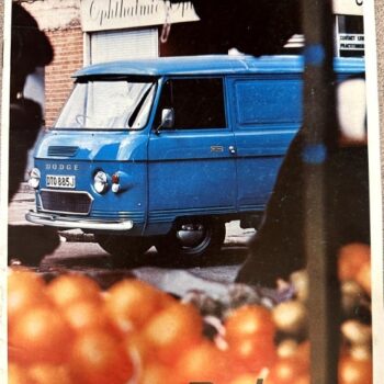 Blue vintage Dodge panel van parked by a market stall, with blurred oranges in the foreground and the Dodge logo in the lower right.
