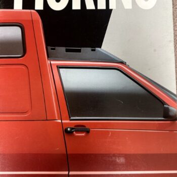 Red Fiat transporter van shown from the side, with a large FIAT sign in the background; visible front door, window, and a 'FIAT Transporter' badge at the bottom.