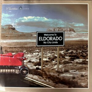 Desert road scene with a red Cadillac tailfin on the left and a black 'Welcome to Eldorado — No City Limits' sign in the foreground.