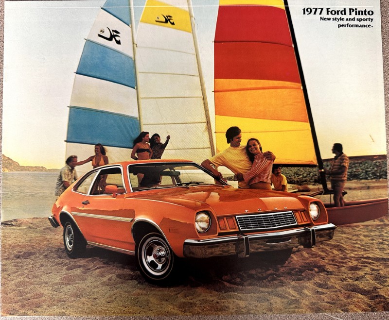 IMG_8877 | Merrick Auto Museum Orange 1977 Ford Pinto parked on a sandy beach with people and colorful sailboats in the background, suggesting a sunny promotional scene.