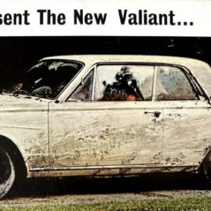 White vintage sedan splattered with mud, parked on grass, with a banner above reading 'We proudly present The New Valiant...'