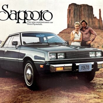 Front-left view of a blue 1978 Sapporo car on a desert road, with a couple standing behind and rock formations in the background.