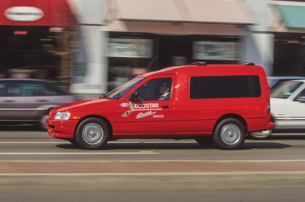 Red EcoStar electric van driving by, with a driver visible in the front seat amid urban storefronts and motion blur.