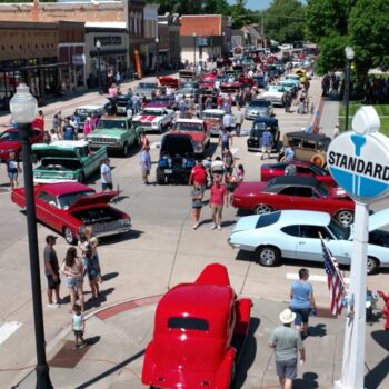 Downtown street filled with colorful classic cars and a crowd at a car show on a sunny day, with a bright red vintage car in the foreground and storefronts in the background.